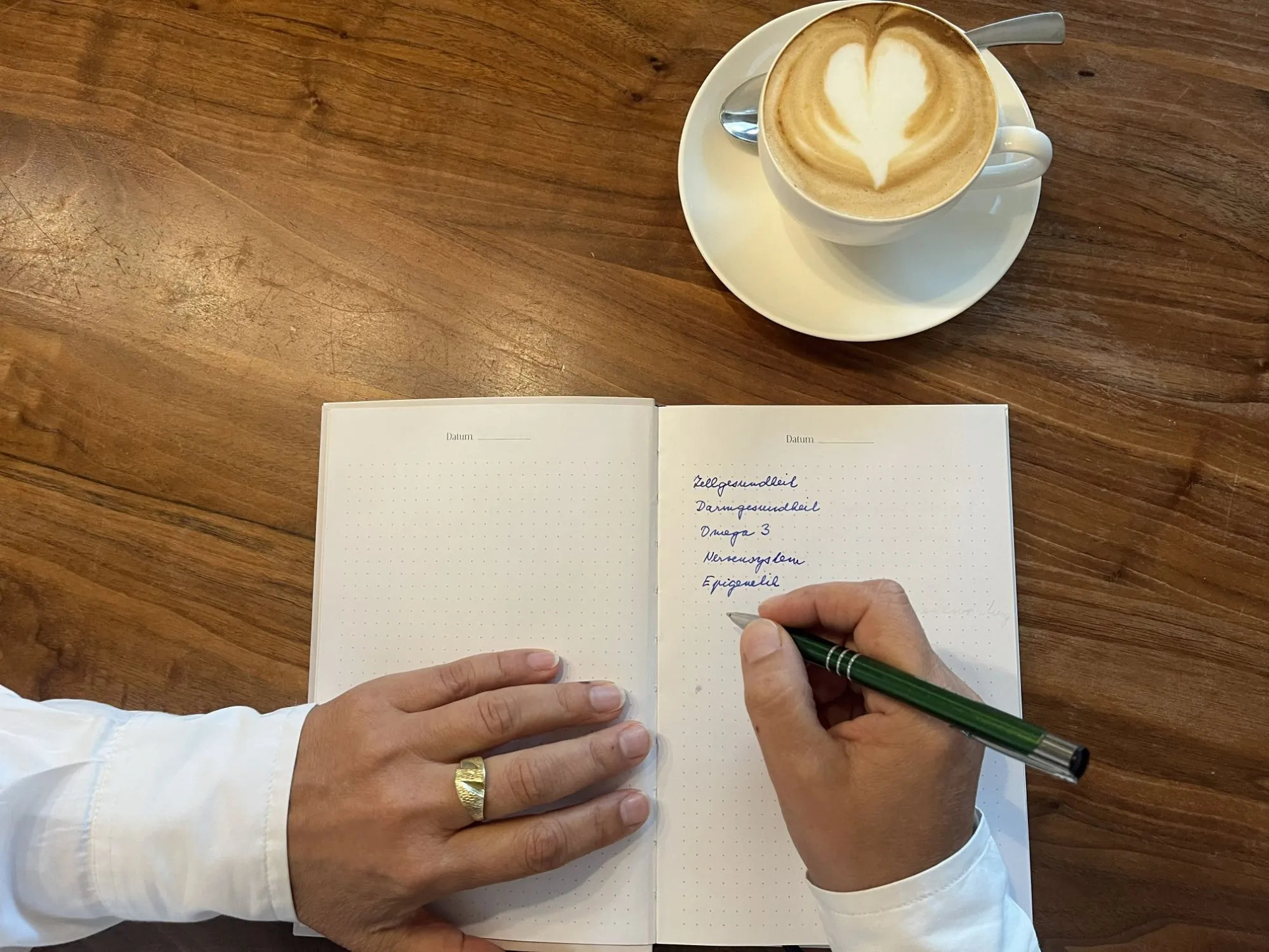Aufgeschlagenes Notizbuch mit handschriftlichen Stichworten — Zellgesundheit, Darmgesundheit, Omega 3, Nervensystem, Epigenetik —, daneben ein Cappuccino mit Milchschaum-Herz